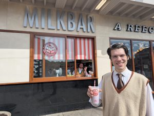Figure holding milkshake, figure in window behind waving.