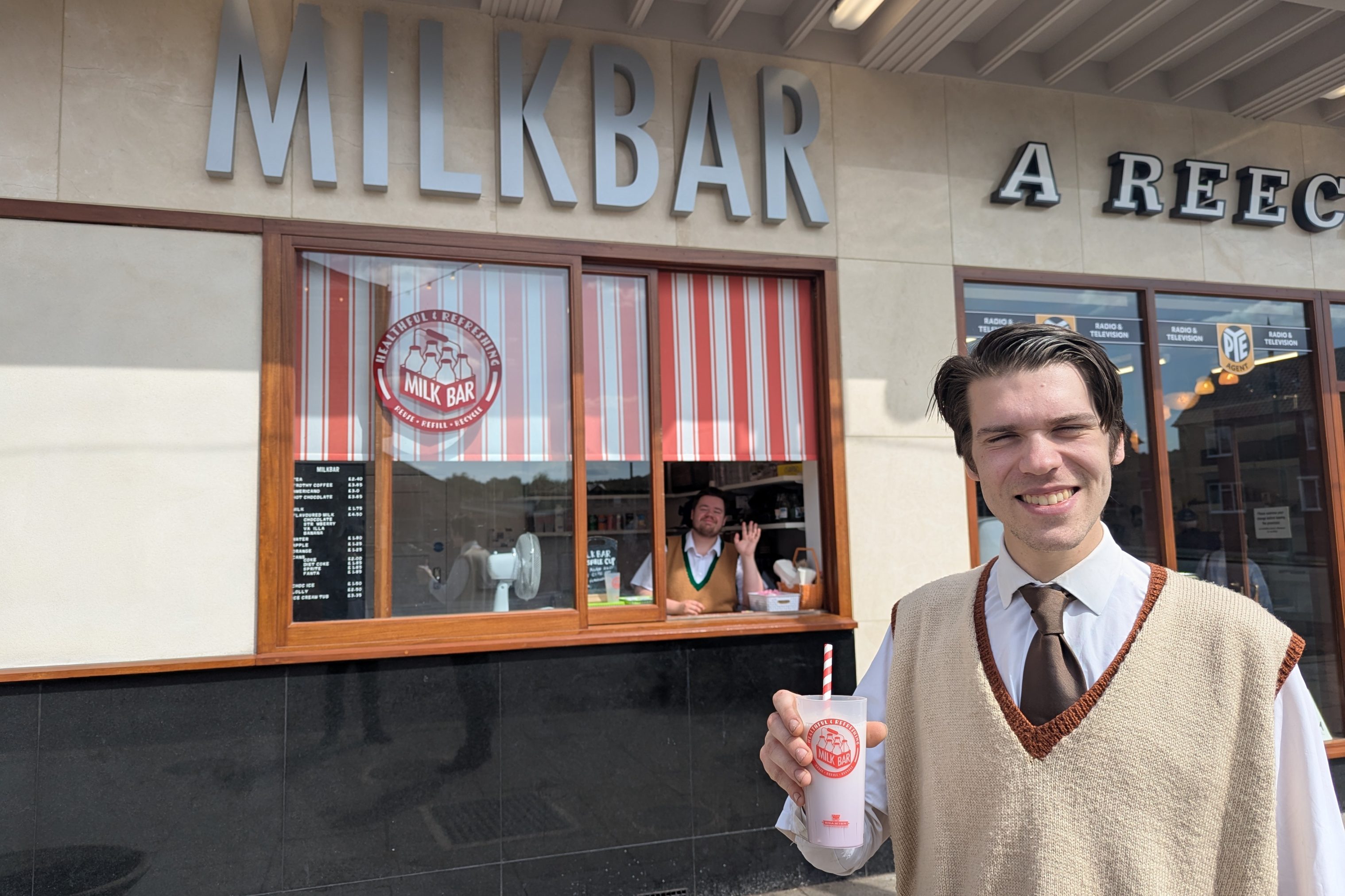 Figure holding milkshake, figure in window behind waving.