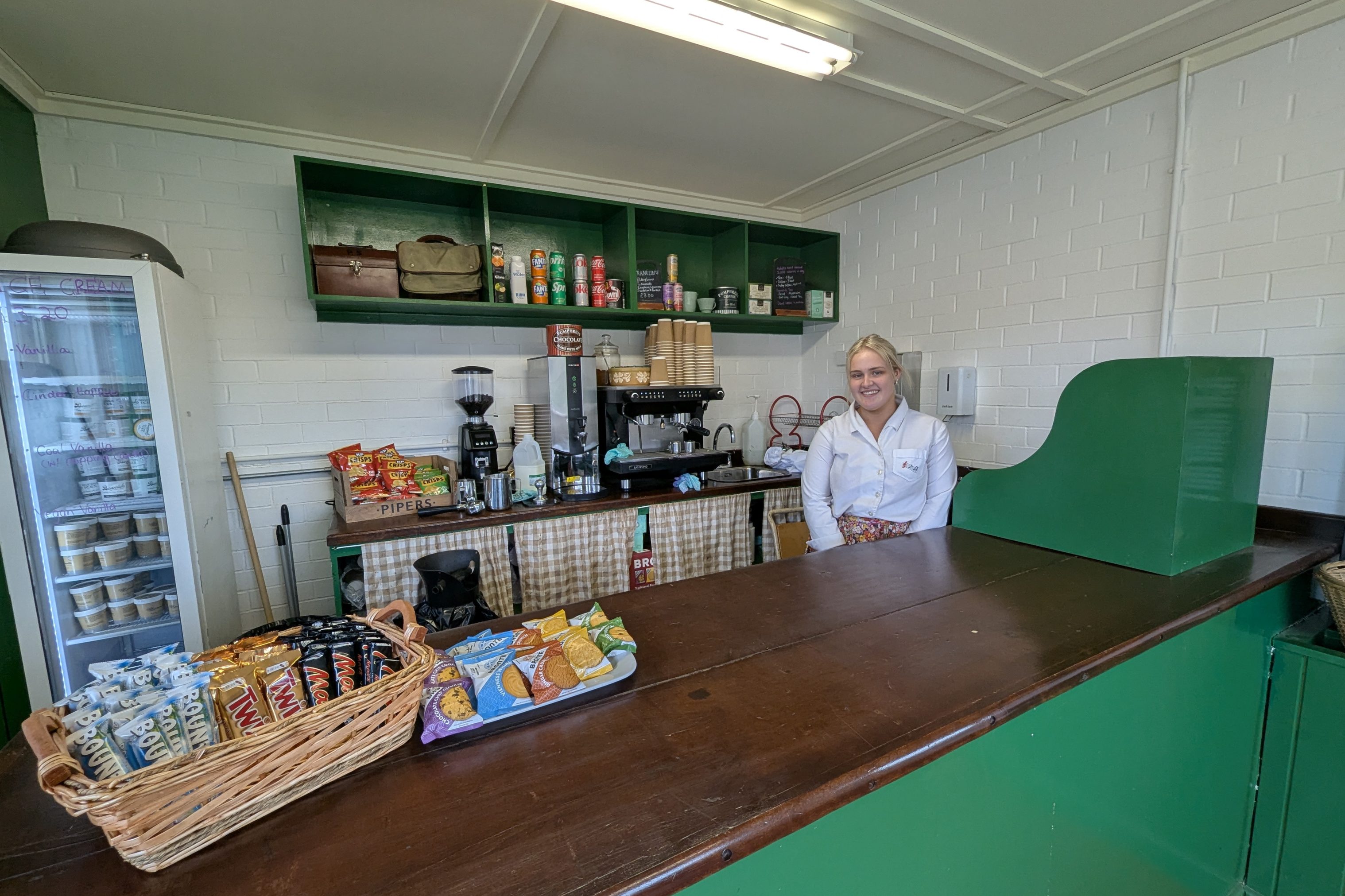 Member of staff behind counter of bowling pavilion. Assortment of snacks on display.