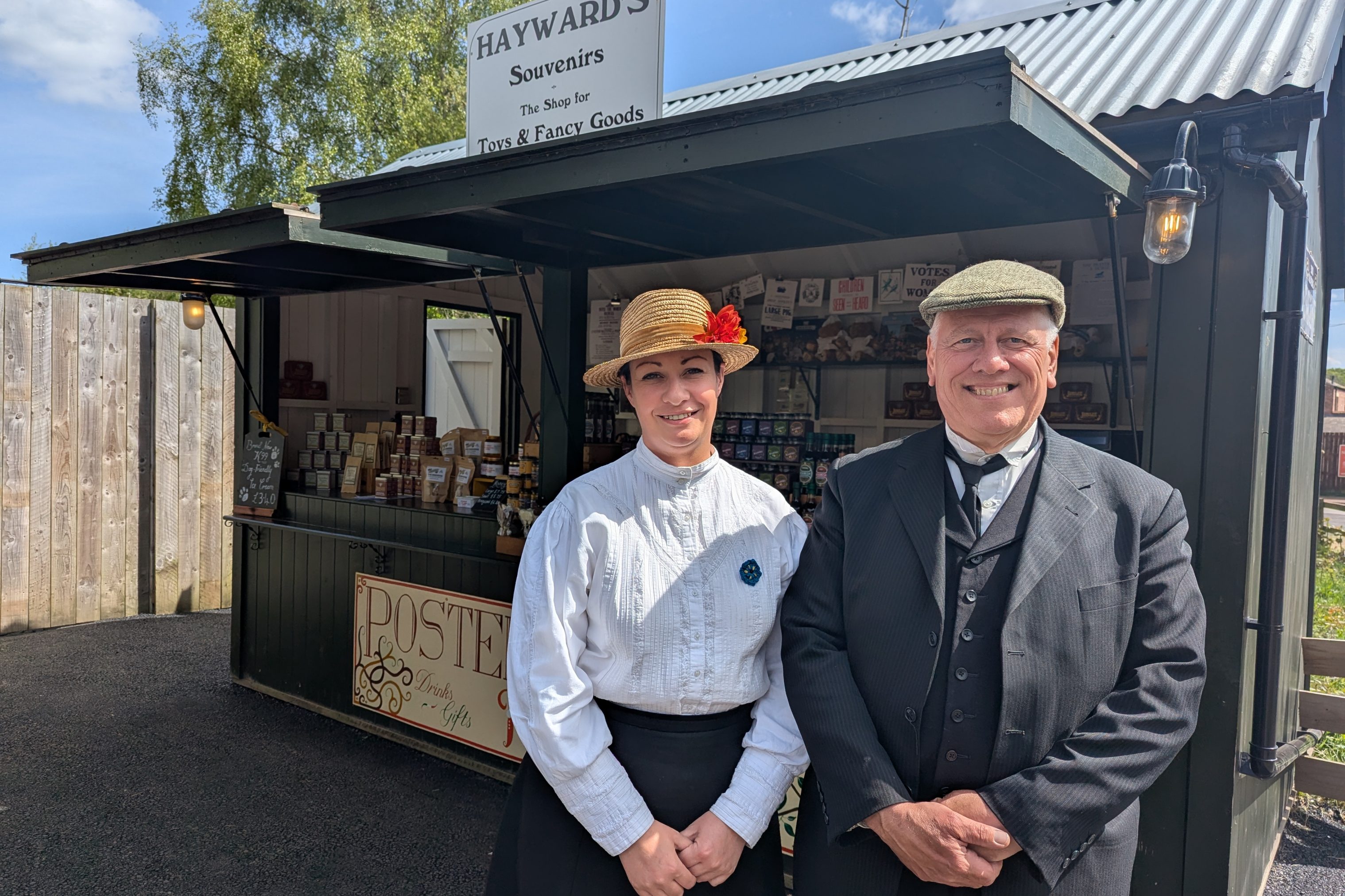Two figures in Edwardian costume, smiling outside town market stall.