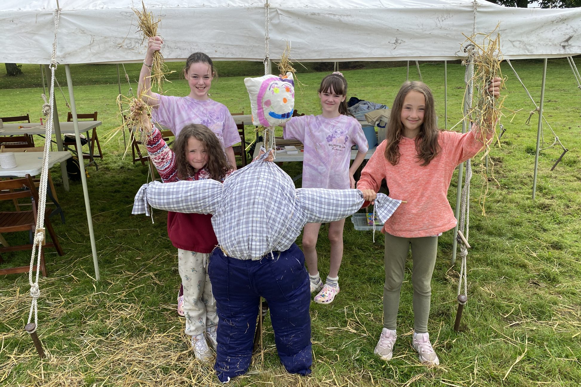 Group of young people holding up hay, next to a scarecrow they've made.