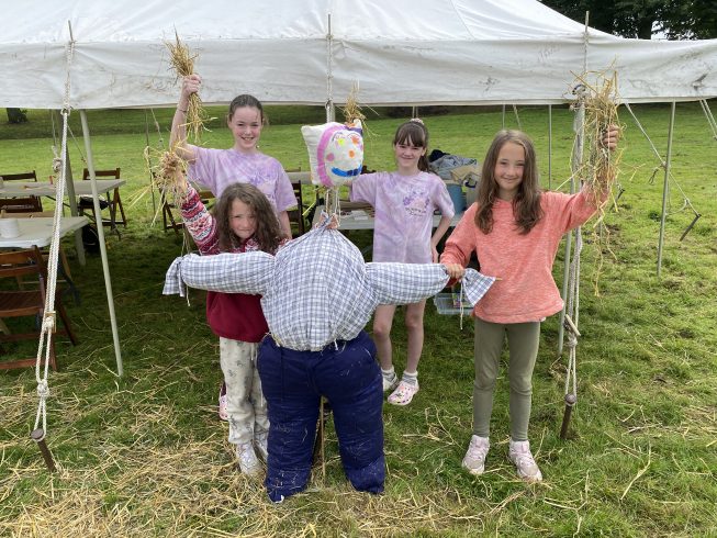 Group of young people holding up hay, next to a scarecrow they've made.