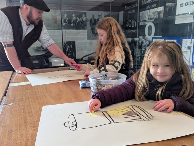 Beamish staff member chatting with two young people drawing a lamp.