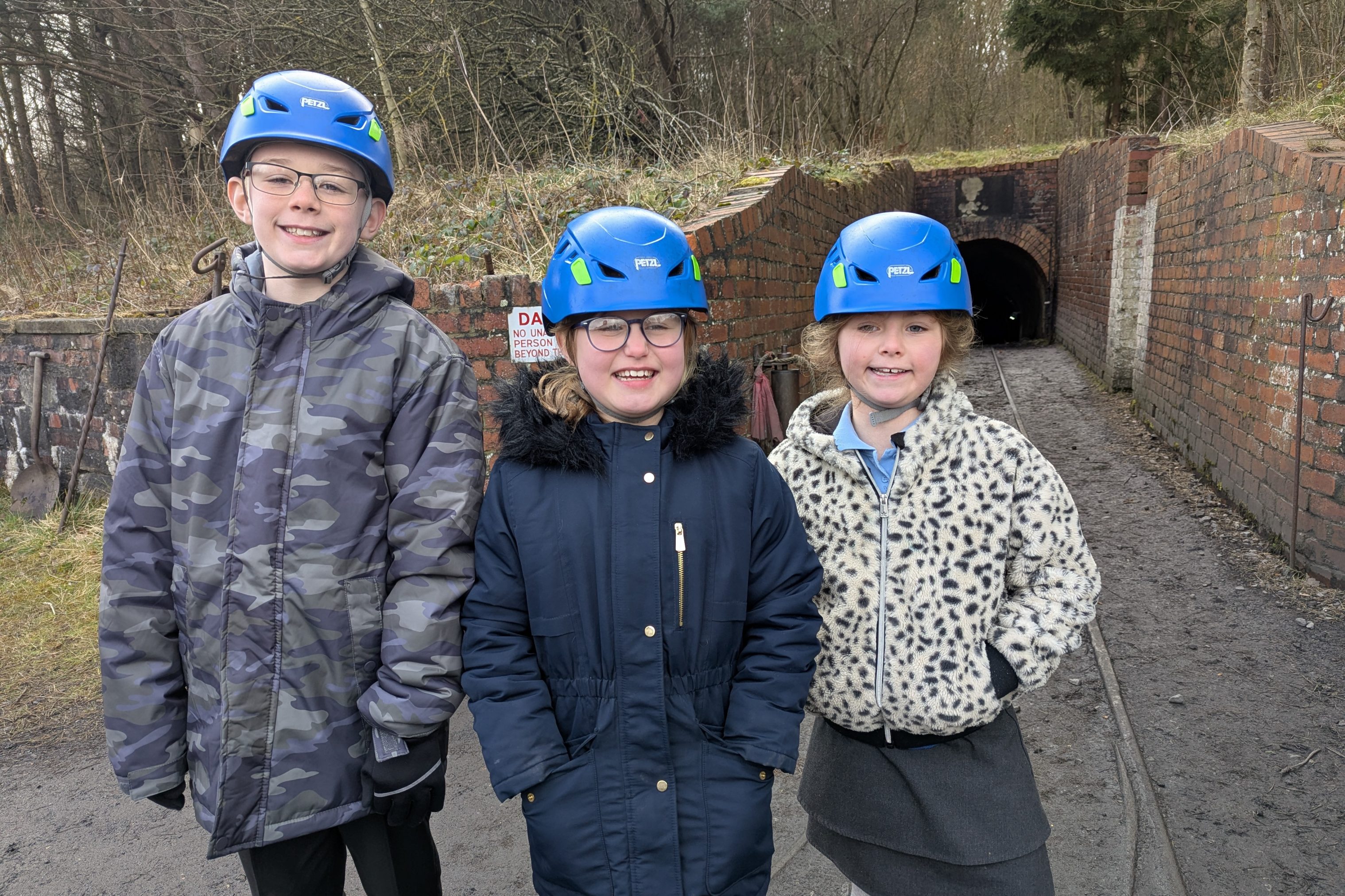 Three young people wearing hard hats, stood in front of drift mine entrance.