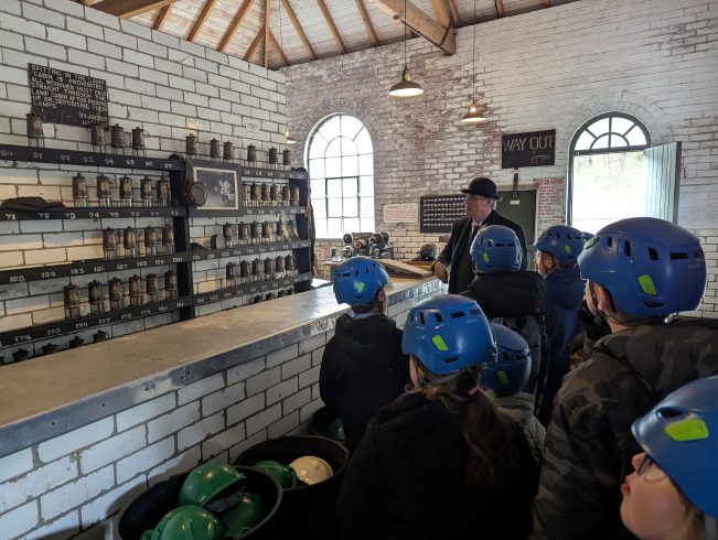 Group of young people wearing hard hats, with staff member looking at pitmen lamps.