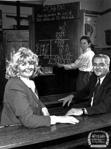 Two figures sat on school bench, with teacher at blackboard behind.