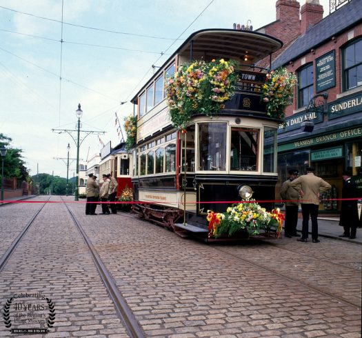 Line of trams covered in flowers on the Town Street, with drivers and conductors stood around.