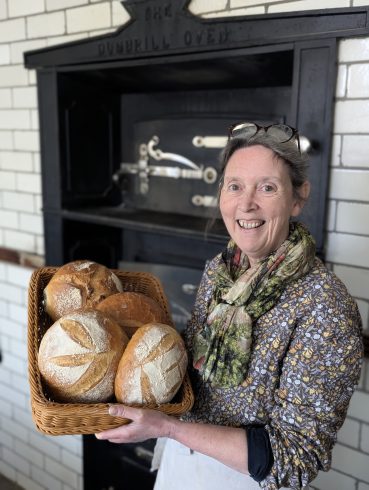 Baker holding a basket full of loaves Infront of a 1900s style oven