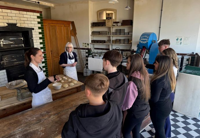 A group of young people stand with their backs to the camera, talking to two people about baking. There is dough on a table as the young people learn the skills needed to bake bread.