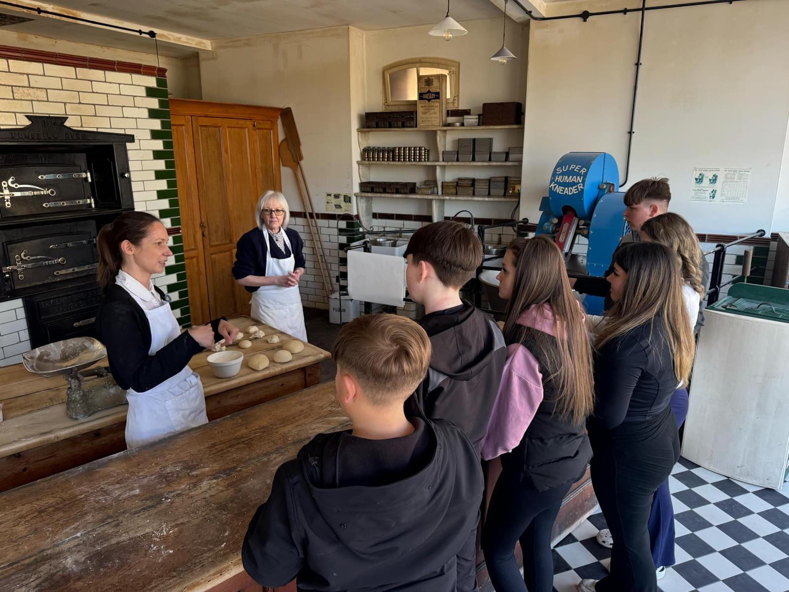 A group of young people stand with their backs to the camera, talking to two people about baking. There is dough on a table as the young people learn the skills needed to bake bread.