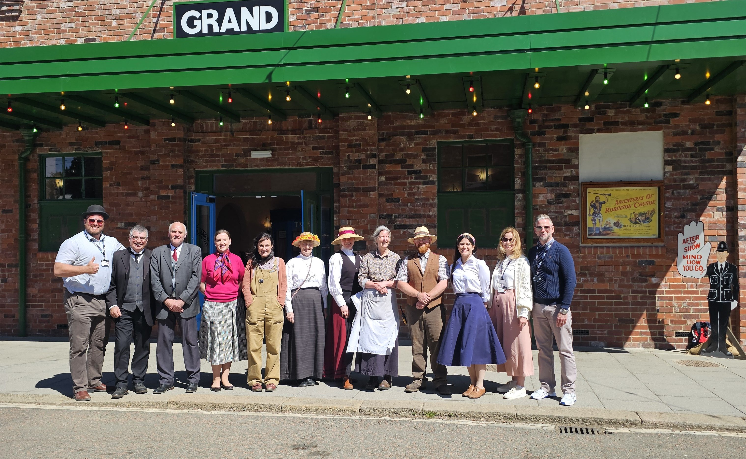 A group of 12 adults stand in row in front of The Grand cinema at Beamish Museum. Many of the people are wearing period costume, representing the 1820s, early 1900s, 1940s and 1950s.