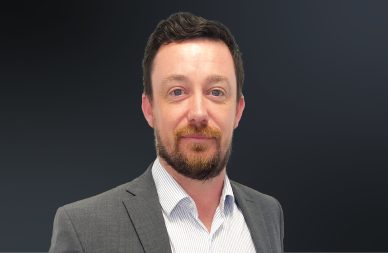 A head shot of Michael Cox wearing a grey suit against a dark background