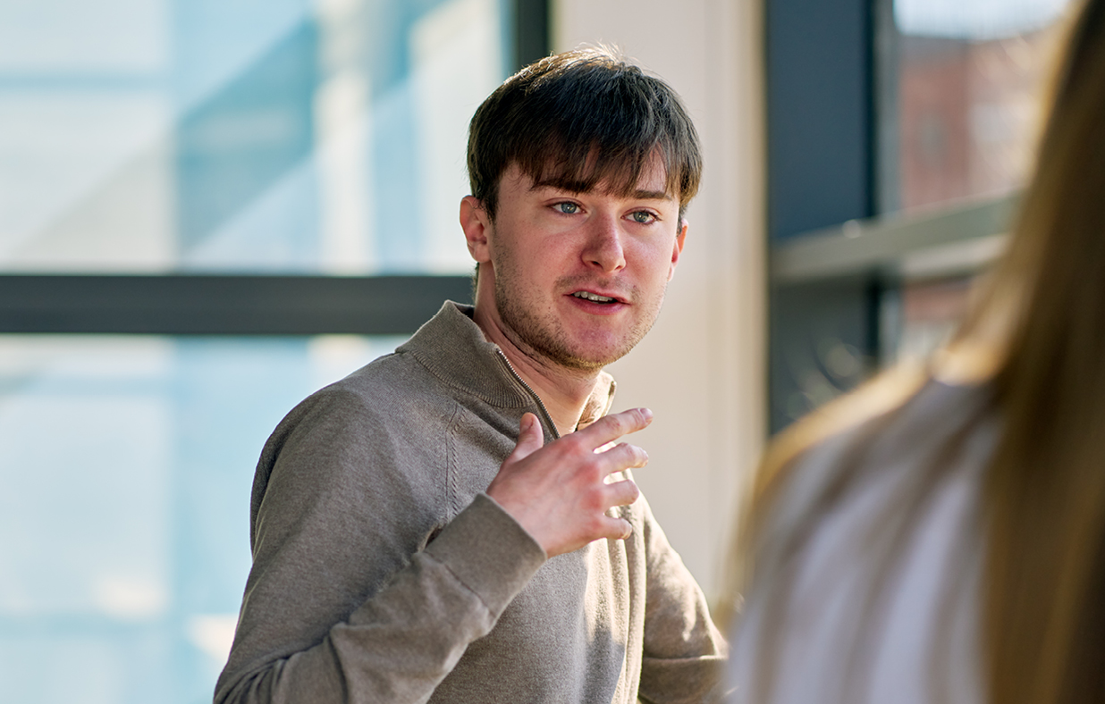 Ben a graduate engineer in conversation with another colleague in an office wearing a light beige jumper.