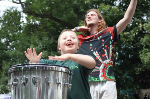 Young boy smiling and drumming outside with adult dancer behind him