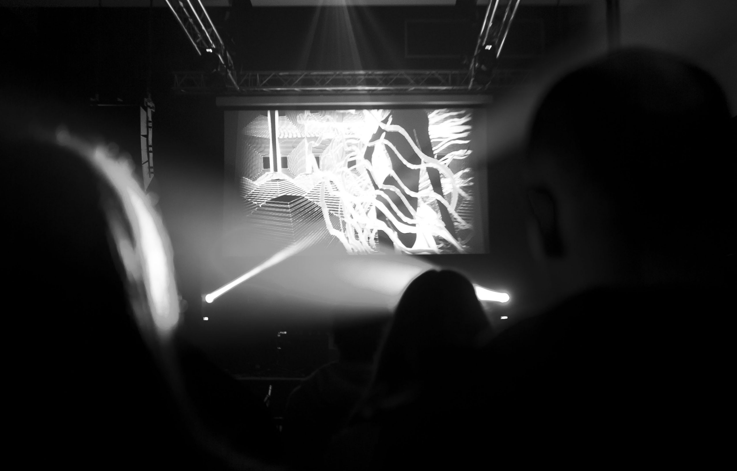 An abstract black-and-white photograph of a performance, with a screen beaming visuals towards an audience.