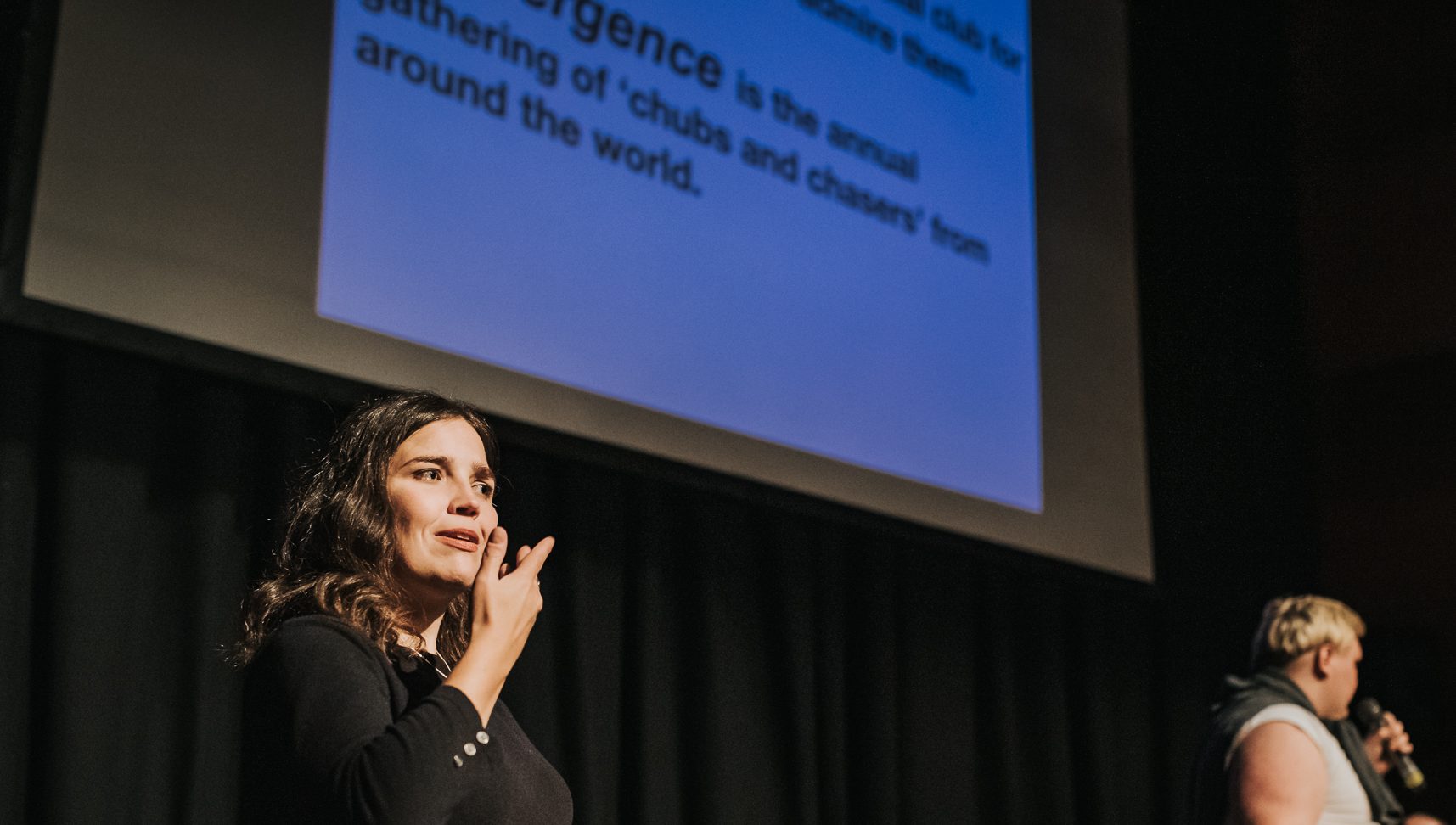 Two people stand on a stage at a film event. The person on the left has brown hair and is signing using BSL. The person on the right speaks into a microphone.