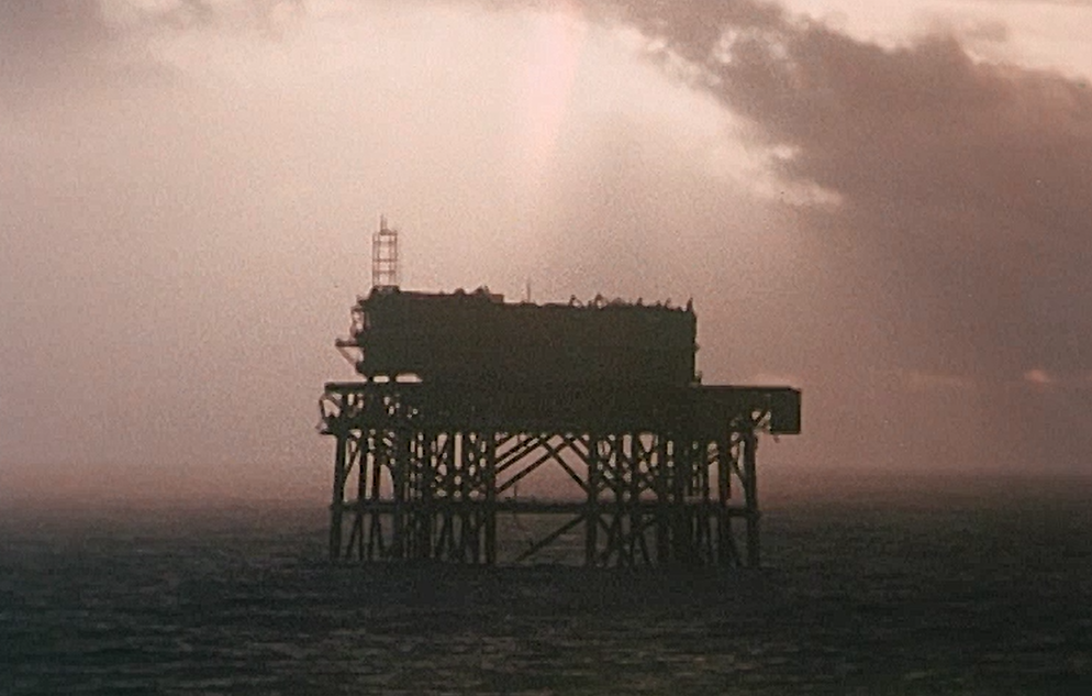 An oil rig is silhouetted against a sepia coloured sky. Still from Living Proof.