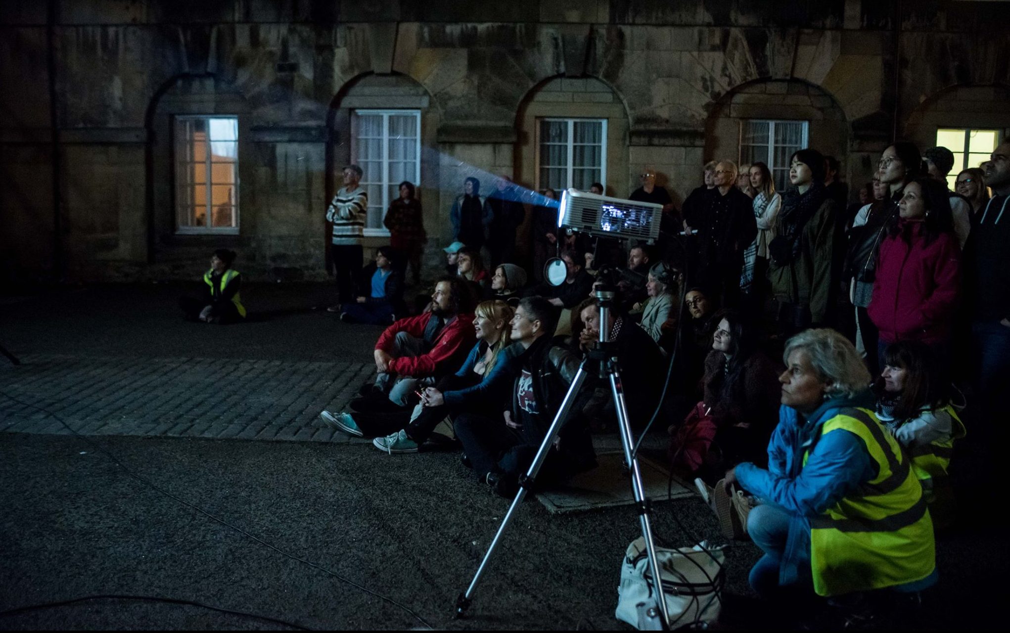 An audience sits on the floor outside, looking towards a projection that's happening off screen. A projector beams light to the left of the image.