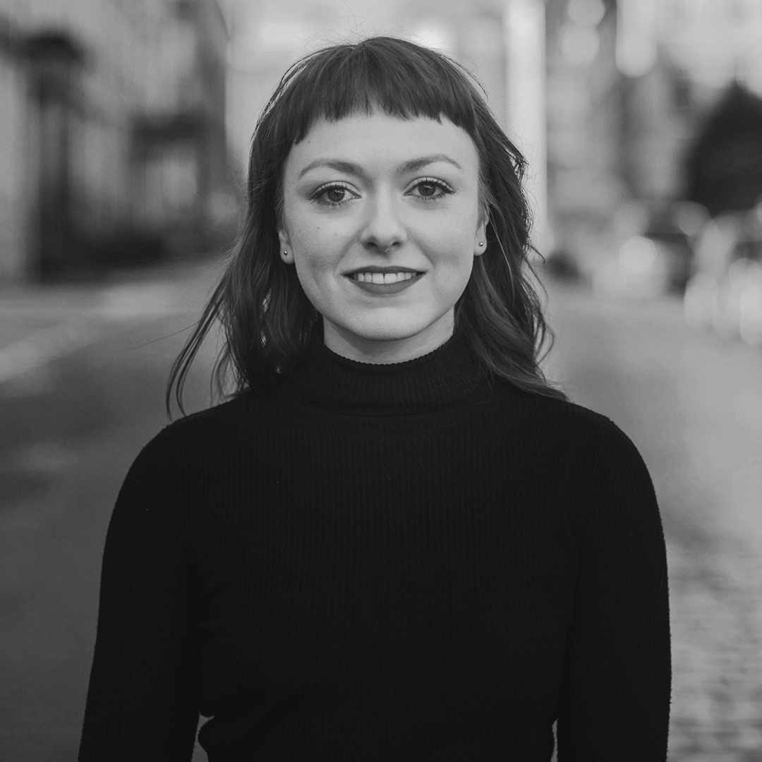 A woman with long brown hair in a black turtleneck stands facing the camera in a street, slightly smiling