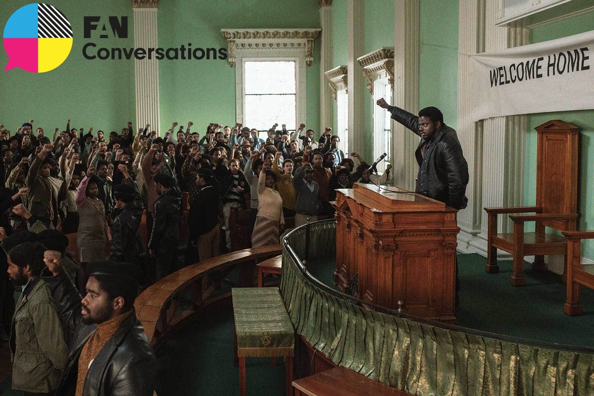 A crowd of Black people stand in a courtroom facing a podium, where a Black man stands with his arm raised, making the Black Power gesture.