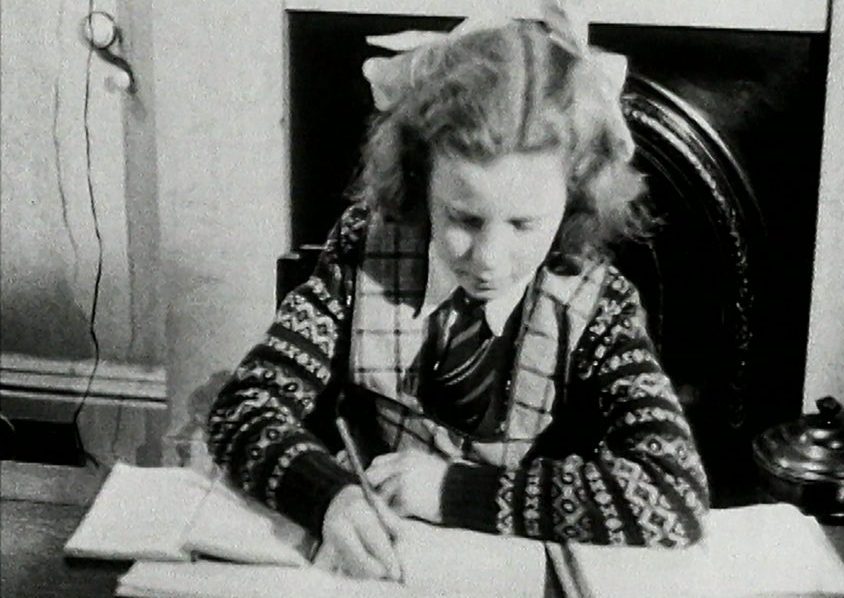 Still from an archive film: a girl sits at a desk, writing in a notebook