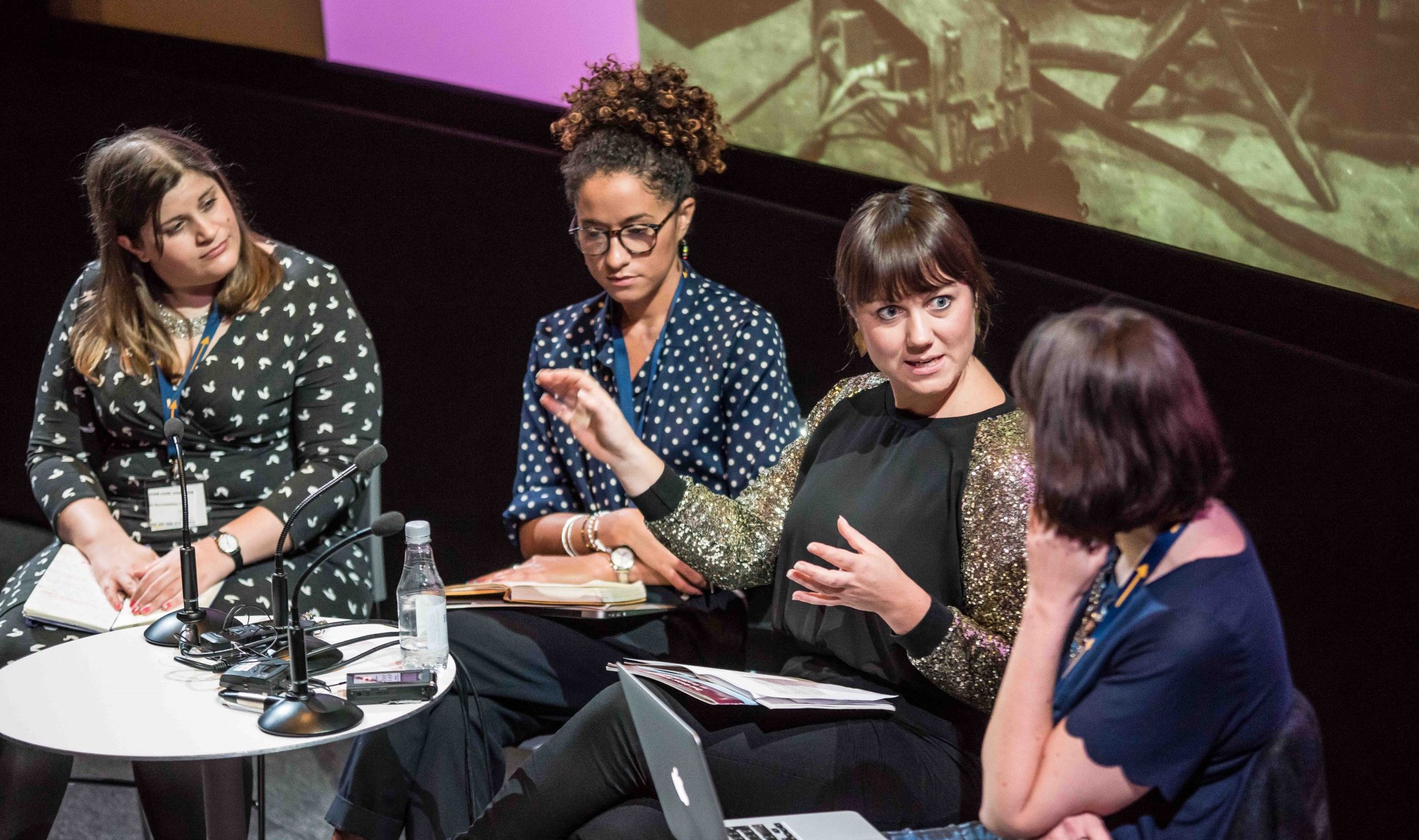 Women sit together on a stage. Three are listening to one speak with animation.