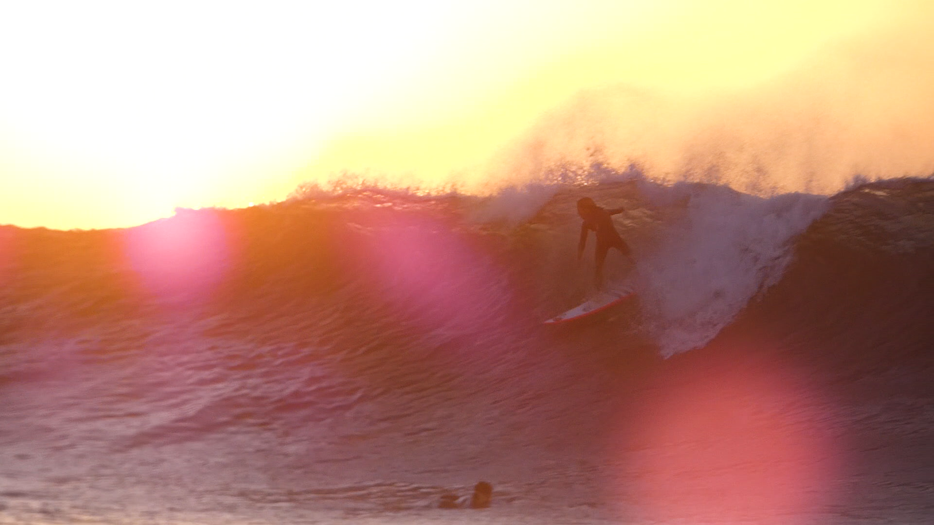 Ride the Wave film still. A young surfer is seen at the crest of a tall wave, bathes in yellow and pink light.