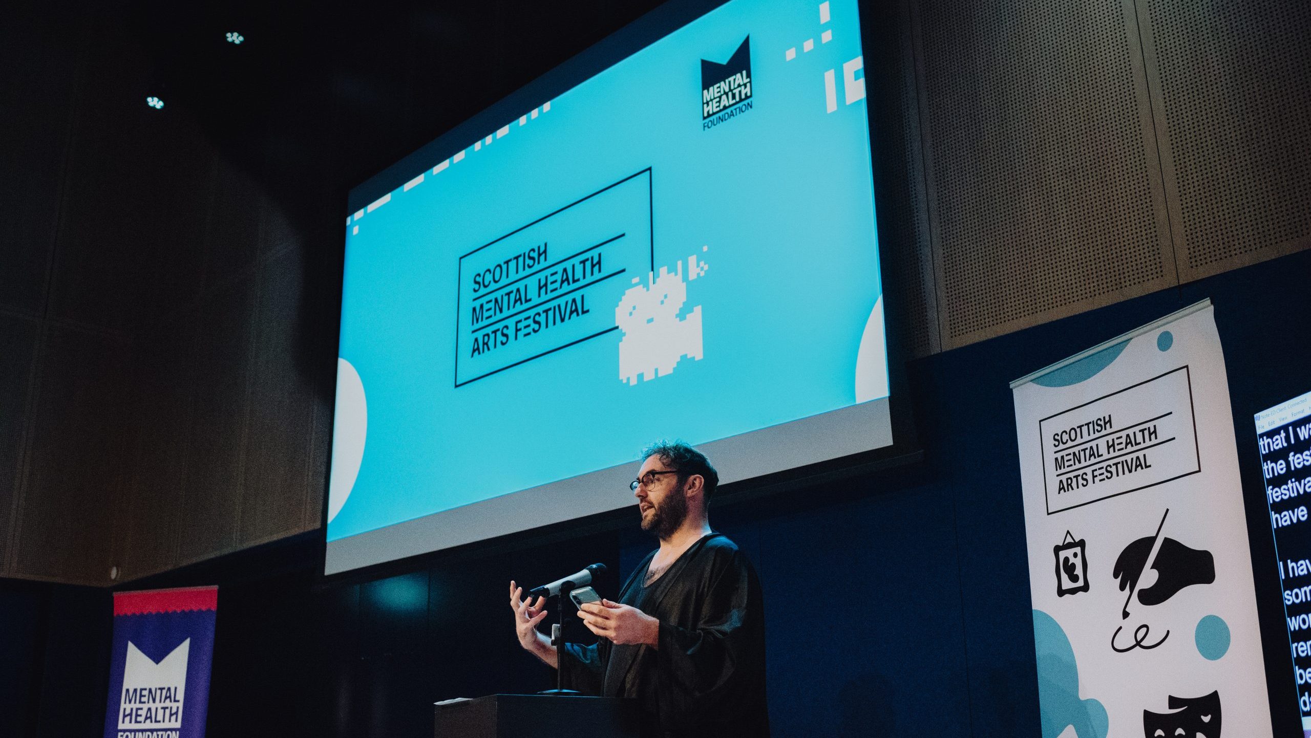 A speaker stands at a podium in a cinema auditorium beneath a large screen displaying Scottish Mental Health Arts Festival branding, with festival banners on either side.