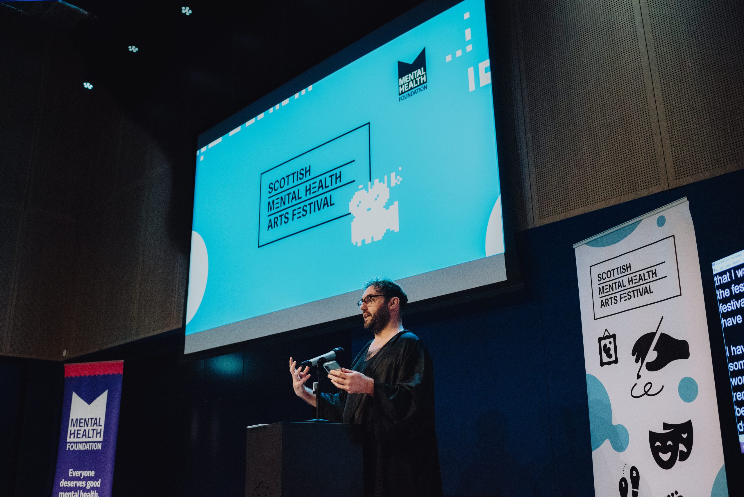 A speaker stands at a podium in a cinema auditorium beneath a large screen displaying Scottish Mental Health Arts Festival branding, with festival banners on either side.