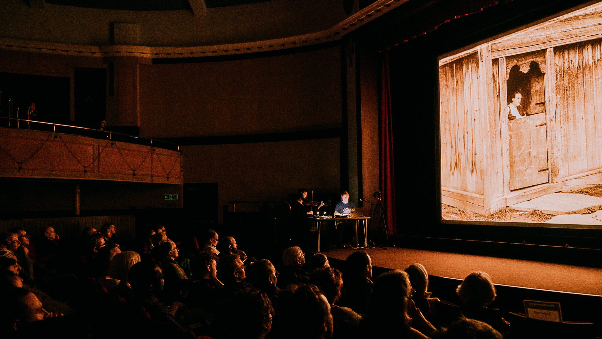 Audience seated in a dark cinema watching a black-and-white film on a large screen, with two people providing live accompaniment at a table beside the screen.