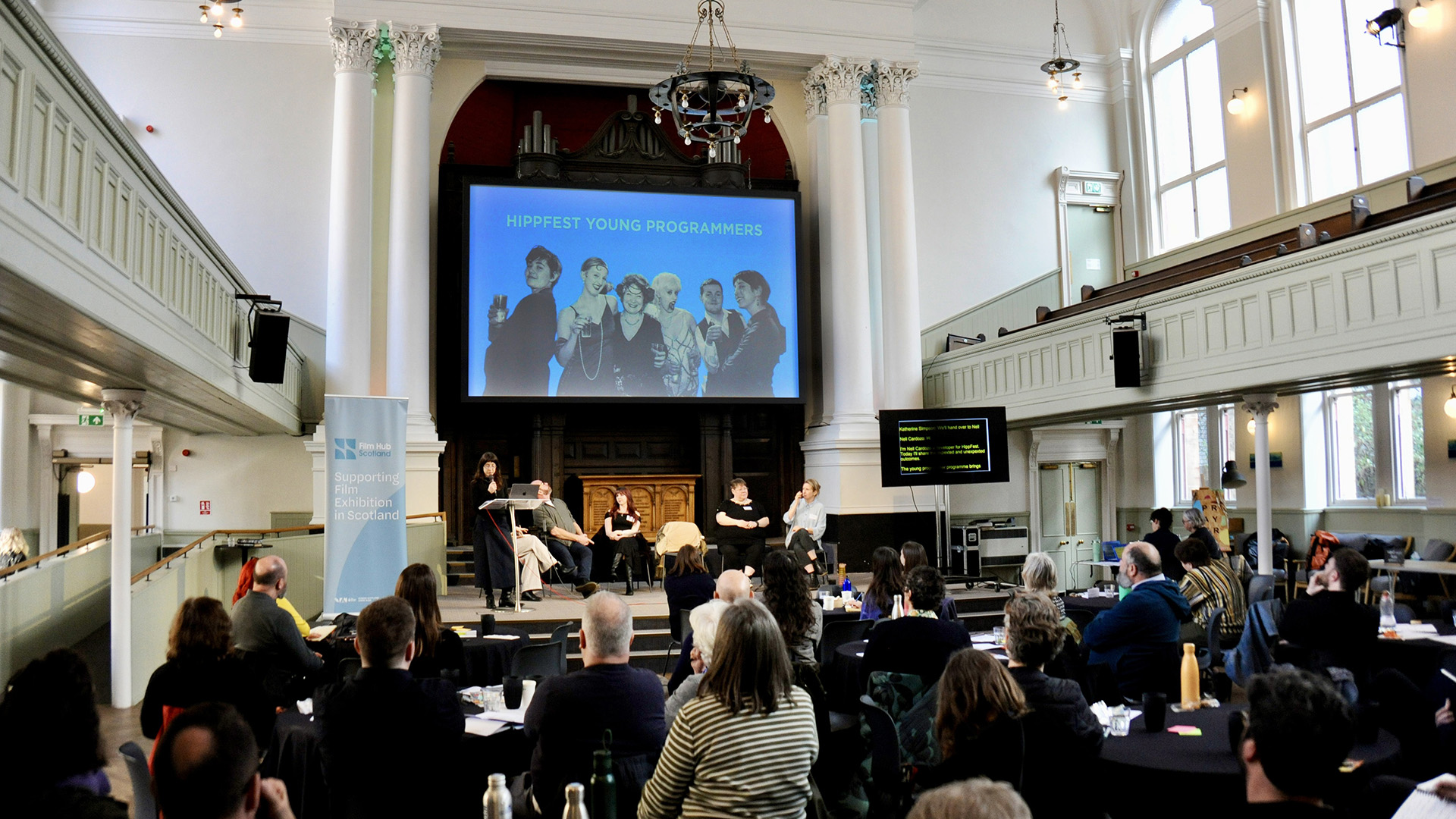 Audience members sit at tables in a large hall watching a panel discussion on stage, with a screen behind the speakers displaying “HippFest Young Programmers.”
