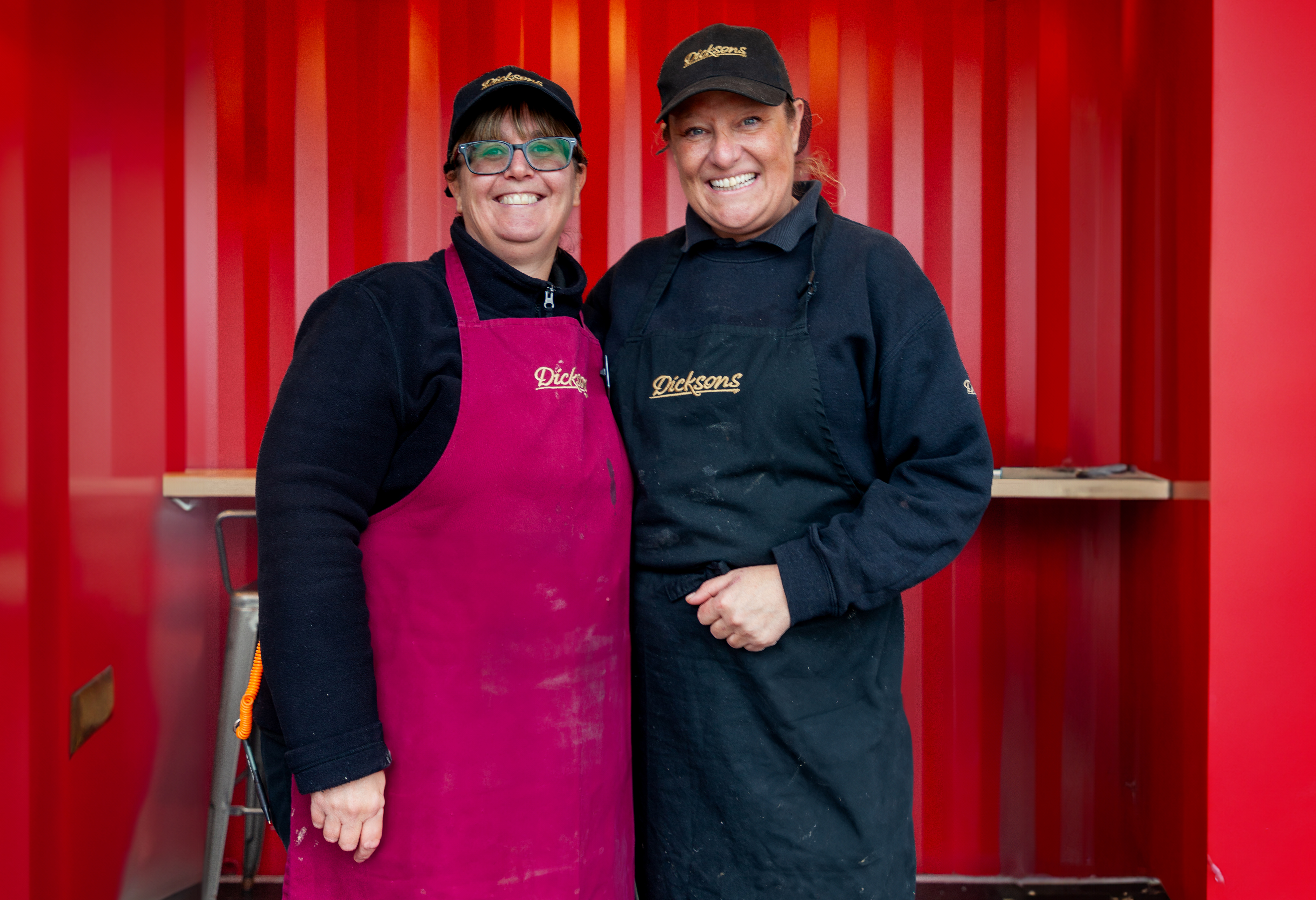 Two woman in aprons smiling against a red background.