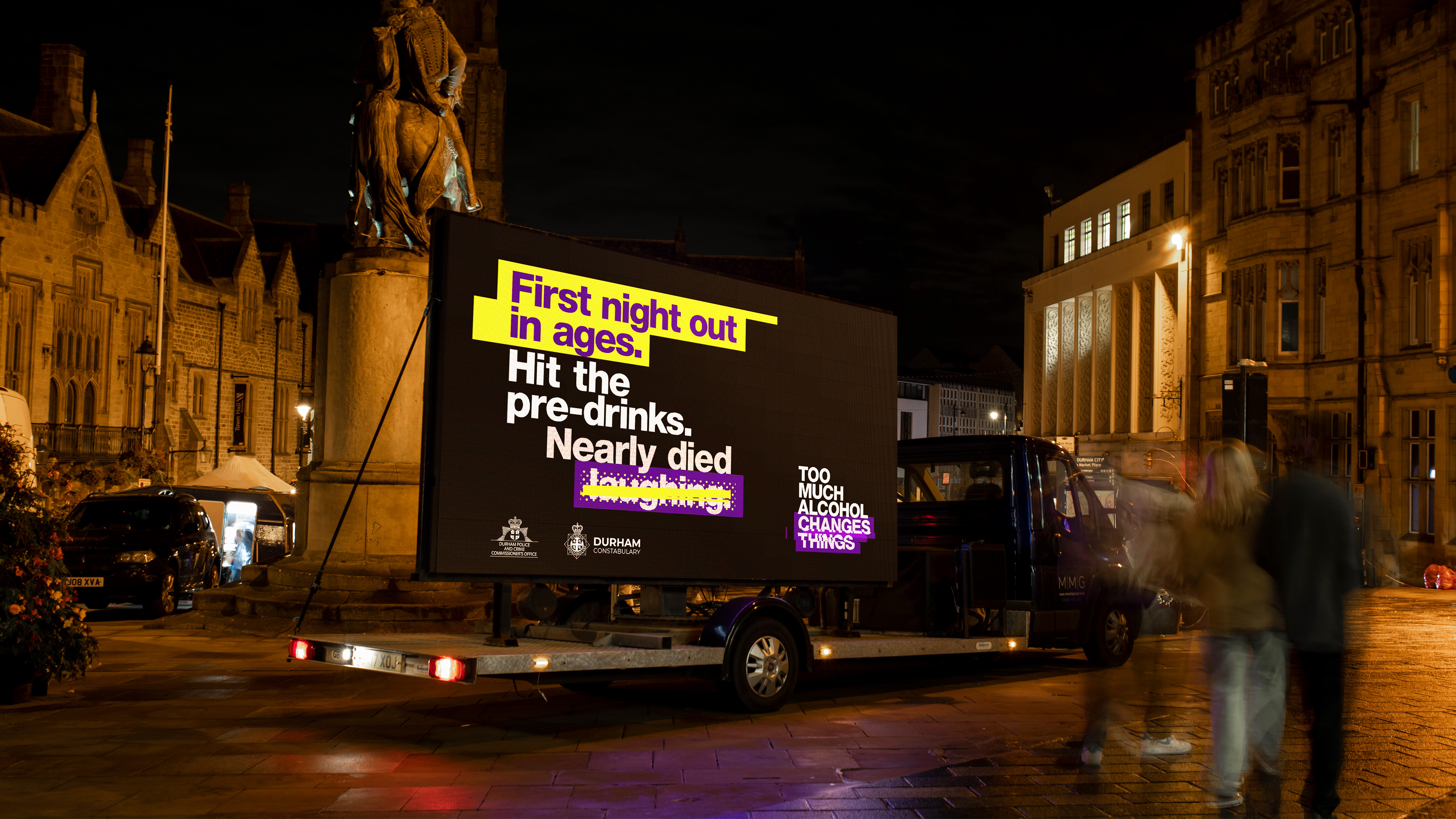 Advertising boarding in middle of a busy town with people passing.