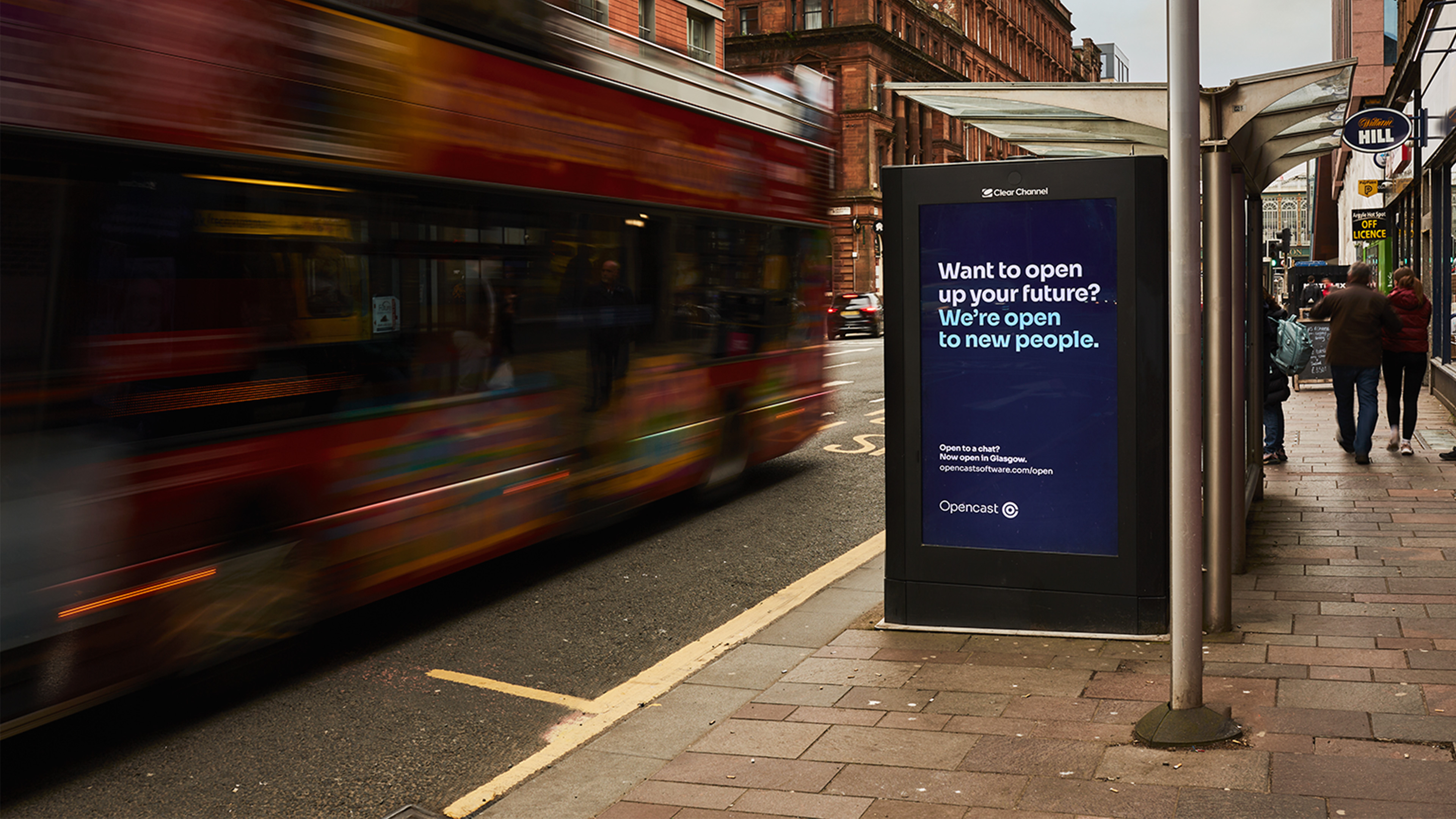 Busy street with people and transport moving past bus shelter with advertising.