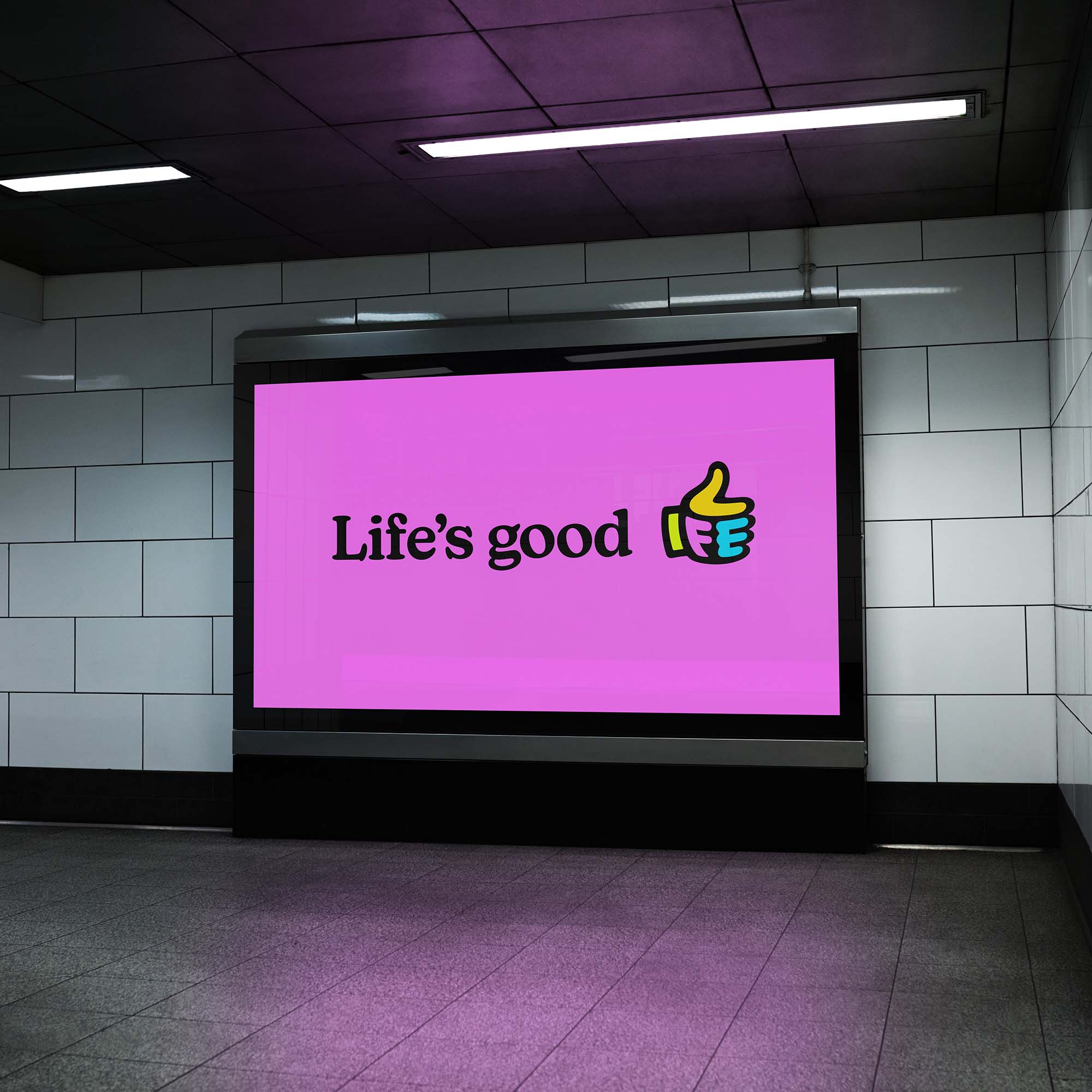 Pink digital ad shell on in a subway. The advert has the text 'Life's good' in bold black text next to the life factory logo which is a thumbs up emoji