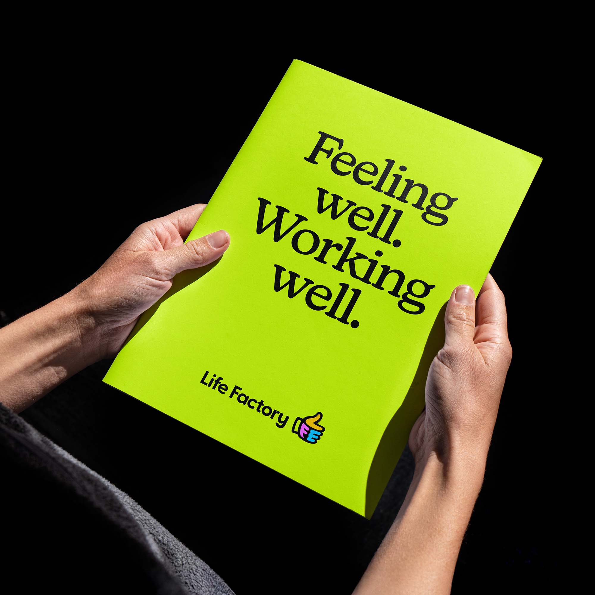 Someone holding a plain bright green brochure against a black background. The brochure cover as big bold lettering with the message 'Feeling well. Working well.