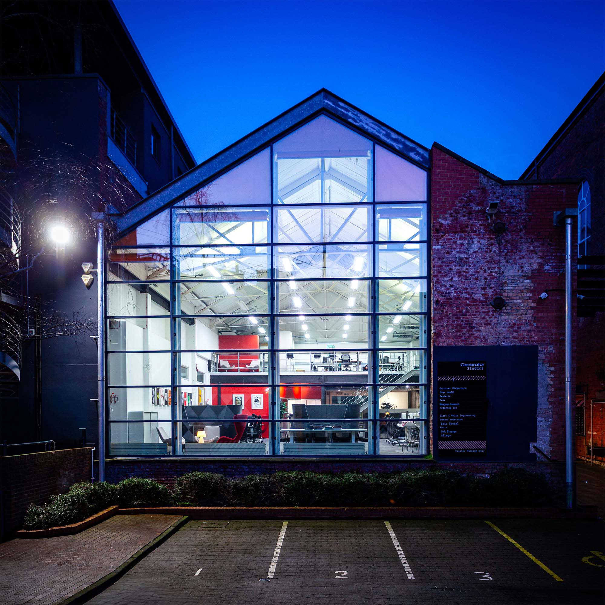 Looking into an old converted warehouse which is has been glazed from floor to ceiling at dusk. Inside is a modern creative workspace.