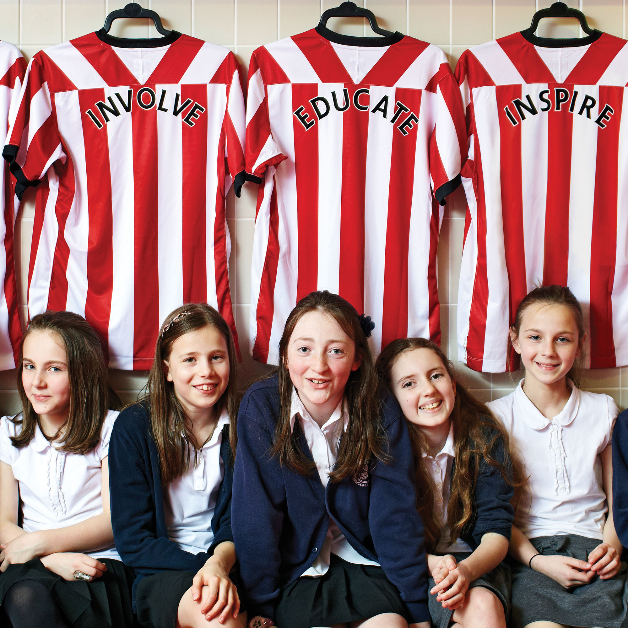 Image of school children sat in front of club strips on the wall of Sunderland AFC changing rooms. The wording Involve, Educate, Inspire is printed on the back of three tops.