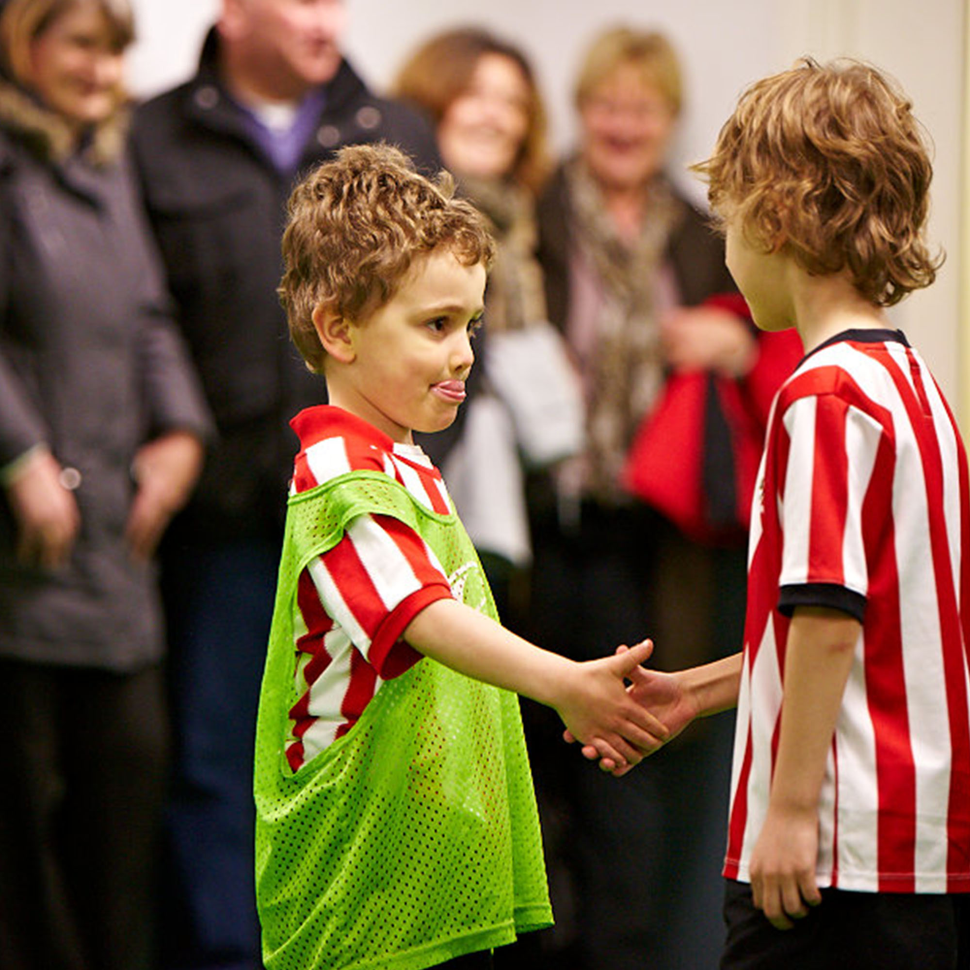 Two children shaking hands on a sports field with the parents looking on in the background.