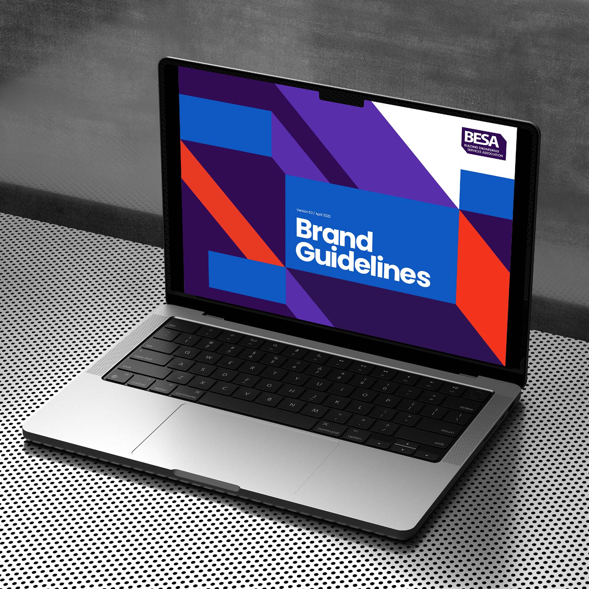 Apple MacBook on a modern steel background. The screen is displaying the front page of the new BESA brand guidelines.
