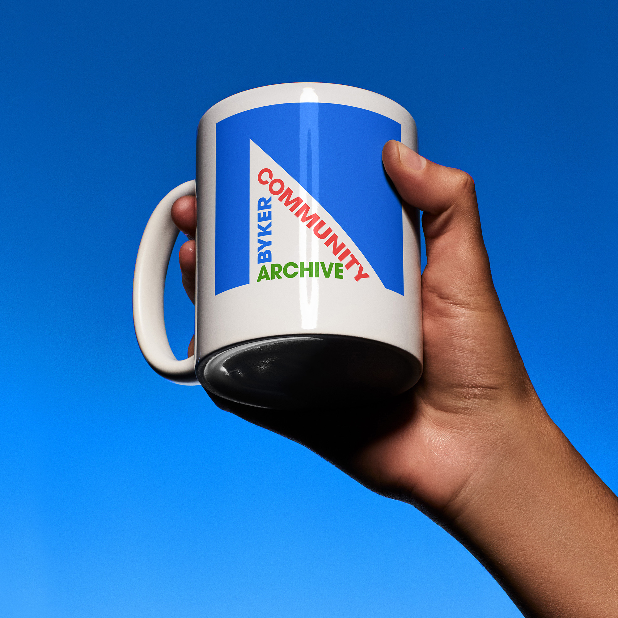 Hand holding a white mug, with the Byker Community Archive logo, against a blue sky background.